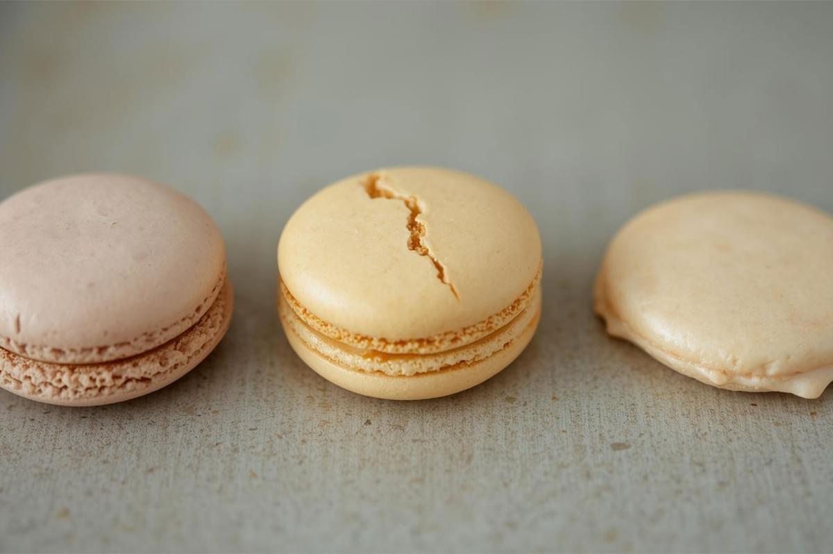 A side-by-side comparison of three macaron shells: a perfect shell on the left, a faulty shell in the center with a prominent crack on top, and a flat, collapsed shell on the right, illustrating baking mistakes.