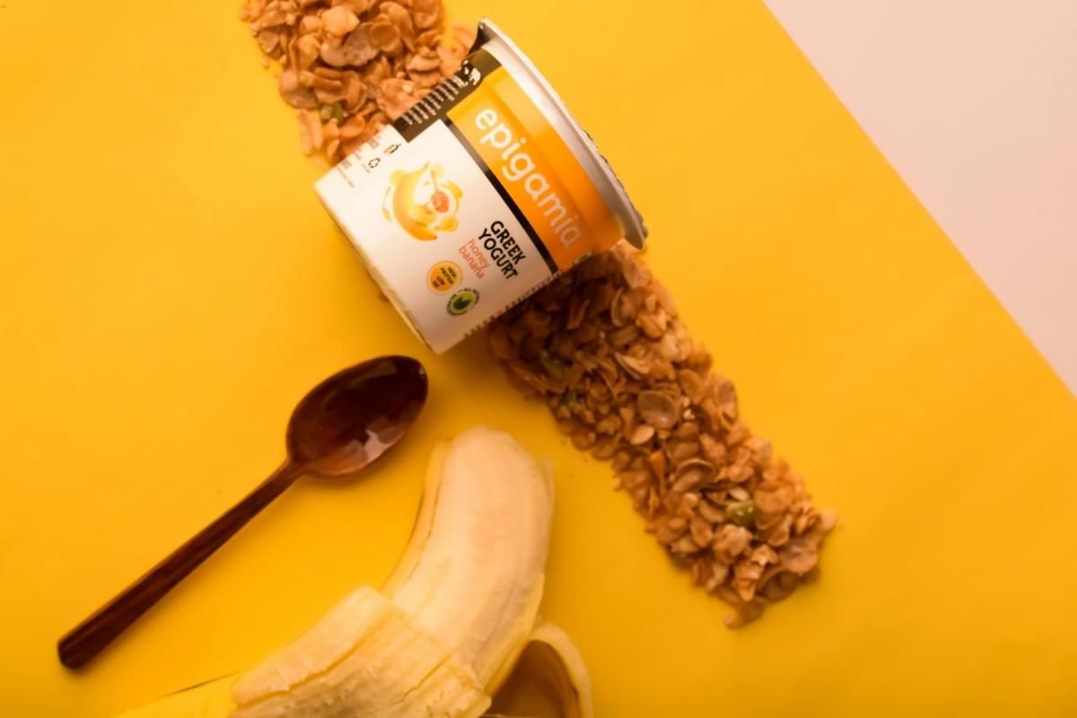 A tub of Epigamia honey banana Greek yogurt is positioned diagonally over a thick line of granola scattered across a bright yellow background. Next to the yogurt, a dark wooden spoon and a partially peeled banana are neatly arranged to complete the breakfast-themed flat lay.