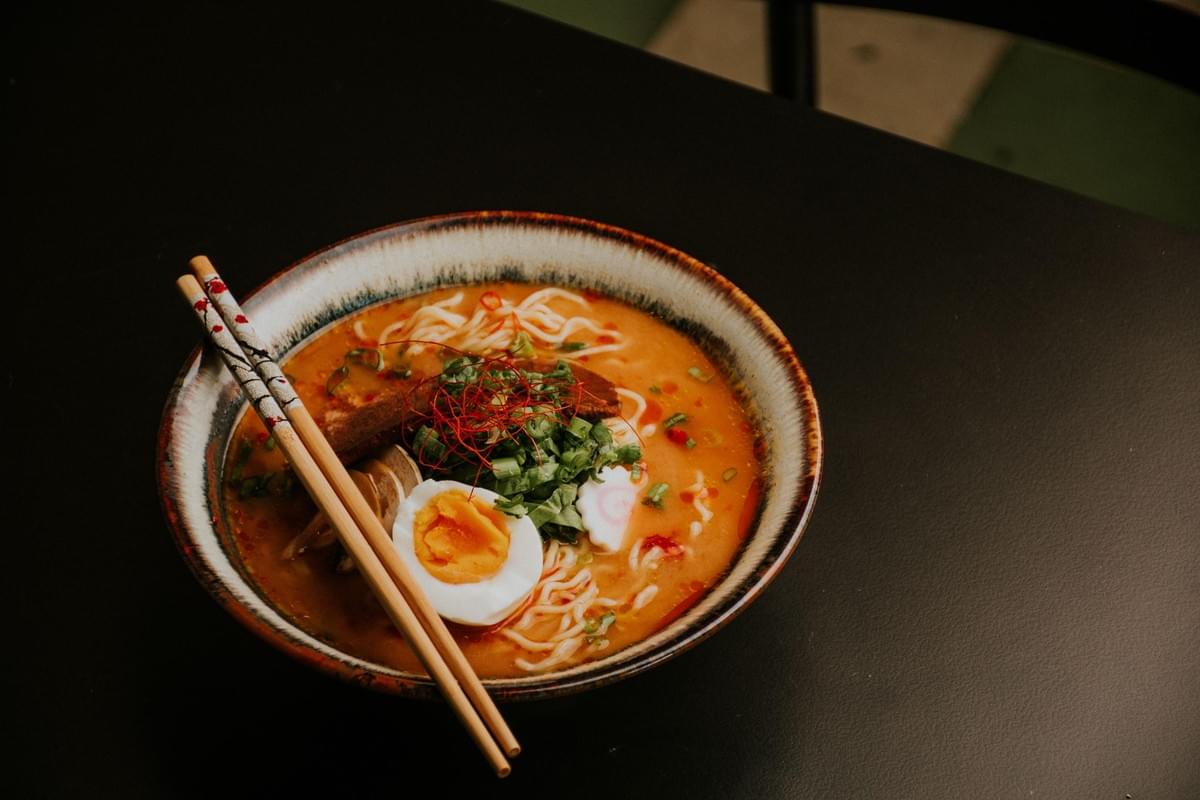 A bowl of ramen with orange broth, noodles, a soft-boiled egg, sliced meat, and green onions sits on a dark table in the image. A pair of chopsticks rests across the rim of the textured ceramic bowl.
