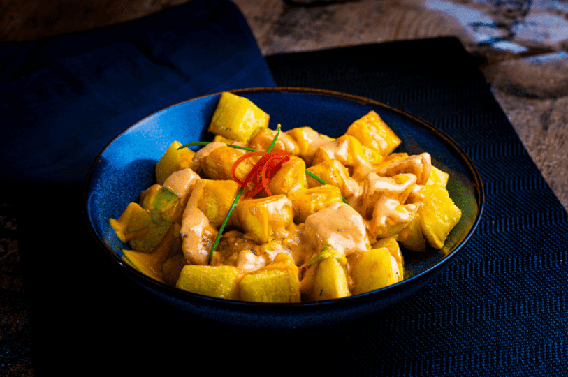 A dark blue ceramic bowl holds a serving of Patatas Bravas, which are golden-yellow, cubed and roasted potatoes. They are generously drizzled with a creamy, pale orange sauce (likely a spicy aioli or 'salsa brava') and possibly a second sauce (less visible, perhaps a tomato-based one) creating a marbled effect. The dish is garnished with thin slices of bright red chili pepper and green chives or herbs. The bowl sits on a dark, textured placemat, with a deep blue cloth napkin visible in the background, all set under dramatic, low-key lighting.