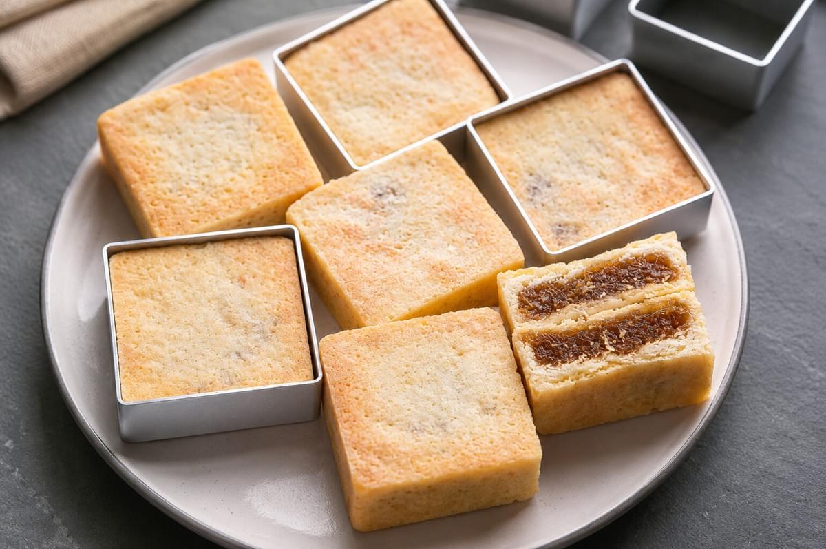 Square pineapple cakes served on a ceramic plate with metal baking molds nearby, showing one cut open to reveal the jam filling.