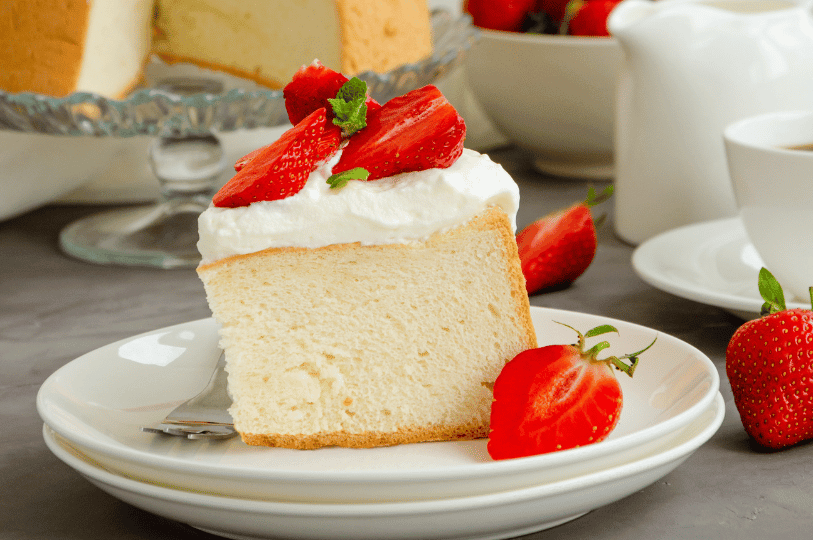 A slice of sponge cake topped with whipped cream, fresh strawberries, and mint sits on a white plate beside a fork—surrounded by berries, a warm drink, and the remaining cake on a stand, evoking a cozy café moment.