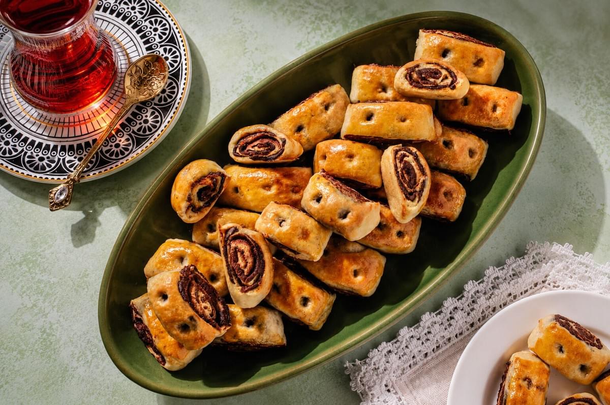Green oval platter filled with golden, date-filled rolled pastries next to a cup of black tea.