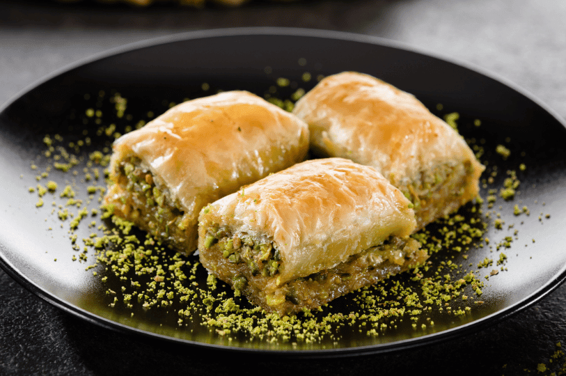 Three rectangular pieces of baklava on a matte black plate, showcasing the glossy, syrupy top layer of pastry and a generous sprinkle of ground nuts.