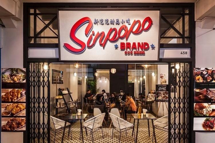 The image showcases the stylish storefront of Sinpopo Brand, featuring a retro-inspired large red logo set against a white perforated background. Through the glass entrance and folding metal gates, a cozy dining area is visible with patrons seated at tables under warm, modern lighting.