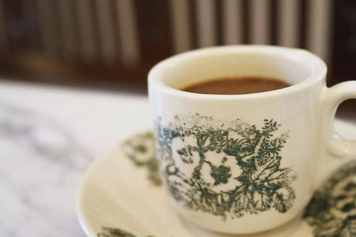 A white ceramic cup filled with coffee sits on a matching saucer. Both have green floral designs, evoking a vintage feel on a marble surface.