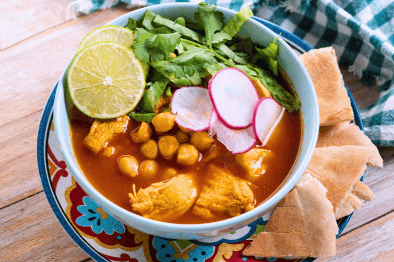 A bright, modern bowl of Pozole Rojo featuring large chunks of chicken or pork and golden hominy, heavily garnished with shredded lettuce, radish, and lime.