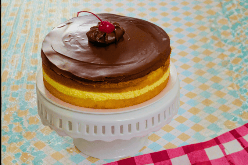 A professional presentation of a Boston Cream Pie on a white pedestal stand, topped with a dollop of chocolate and a bright red maraschino cherry against a checkered pastel background.