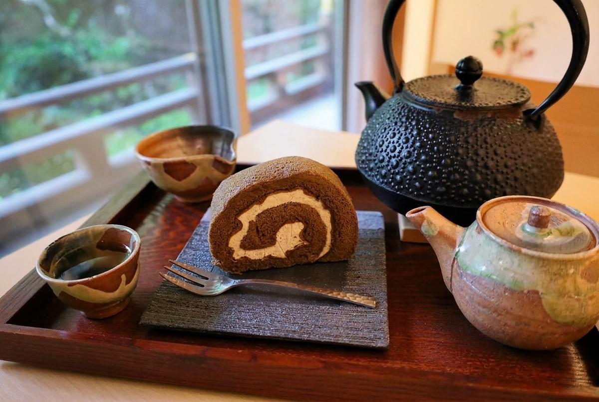 Slice of chocolate roll cake served on a slate plate next to traditional teapots and teacups on a wooden tray.