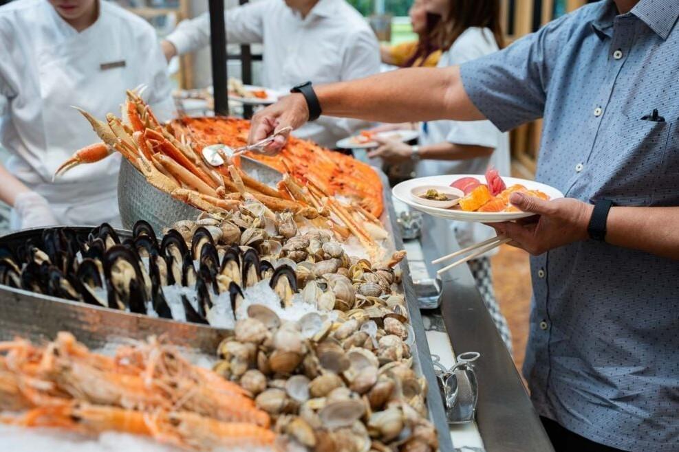 A diner fills a plate with sushi and sashimi while using tongs to select from a lavish seafood buffet spread on ice. The buffet features an abundant selection of fresh seafood, including piles of prawns, clams, mussels, and snow crab legs.