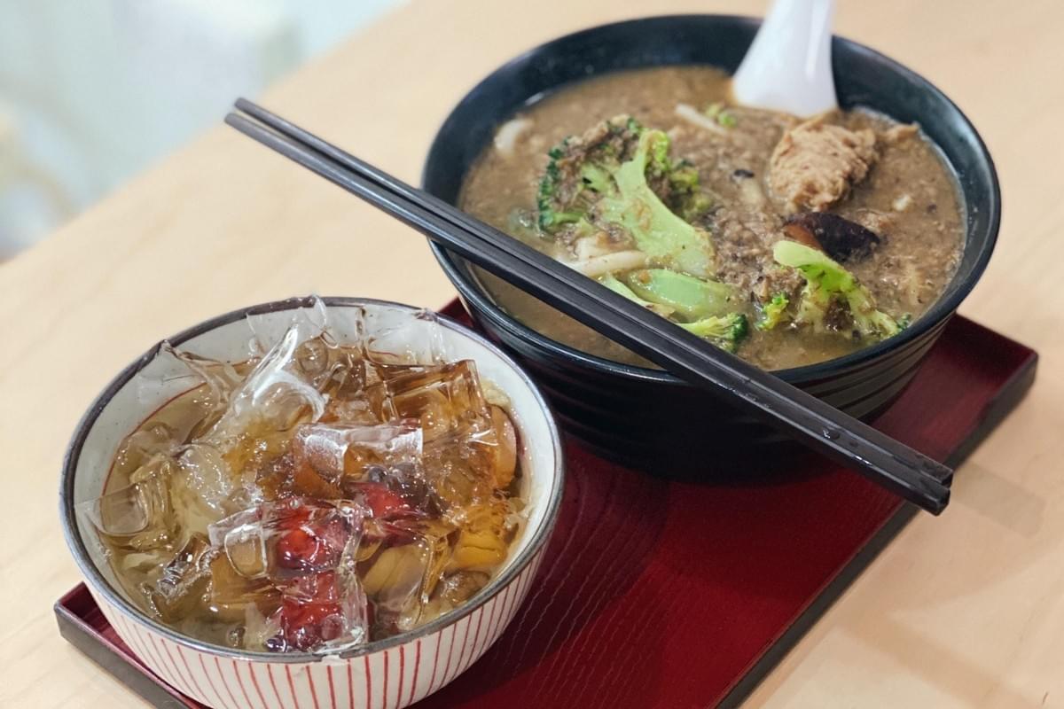 A large black bowl filled with a hearty noodle soup featuring broccoli and mushrooms sits on a red tray alongside a pair of dark chopsticks. Next to it, a smaller striped bowl contains a translucent jelly dessert topped with ice and red dates.