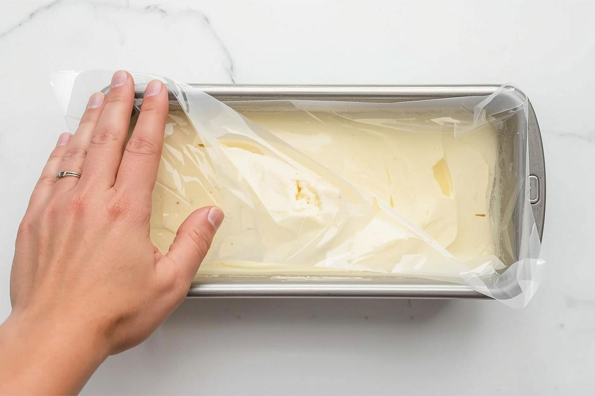An overhead view of a hand covering a stainless steel loaf pan filled with liquid ice cream base with a sheet of plastic wrap before freezing.