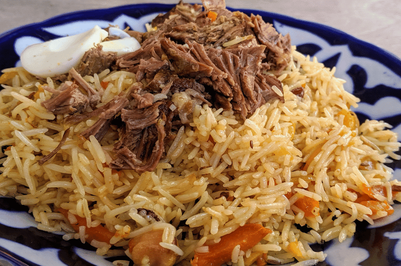 A close-up of Uzbek plov on a decorative plate, highlighting long-grain rice, visible cumin seeds, cooked carrots, and a generous topping of tender, shredded meat.