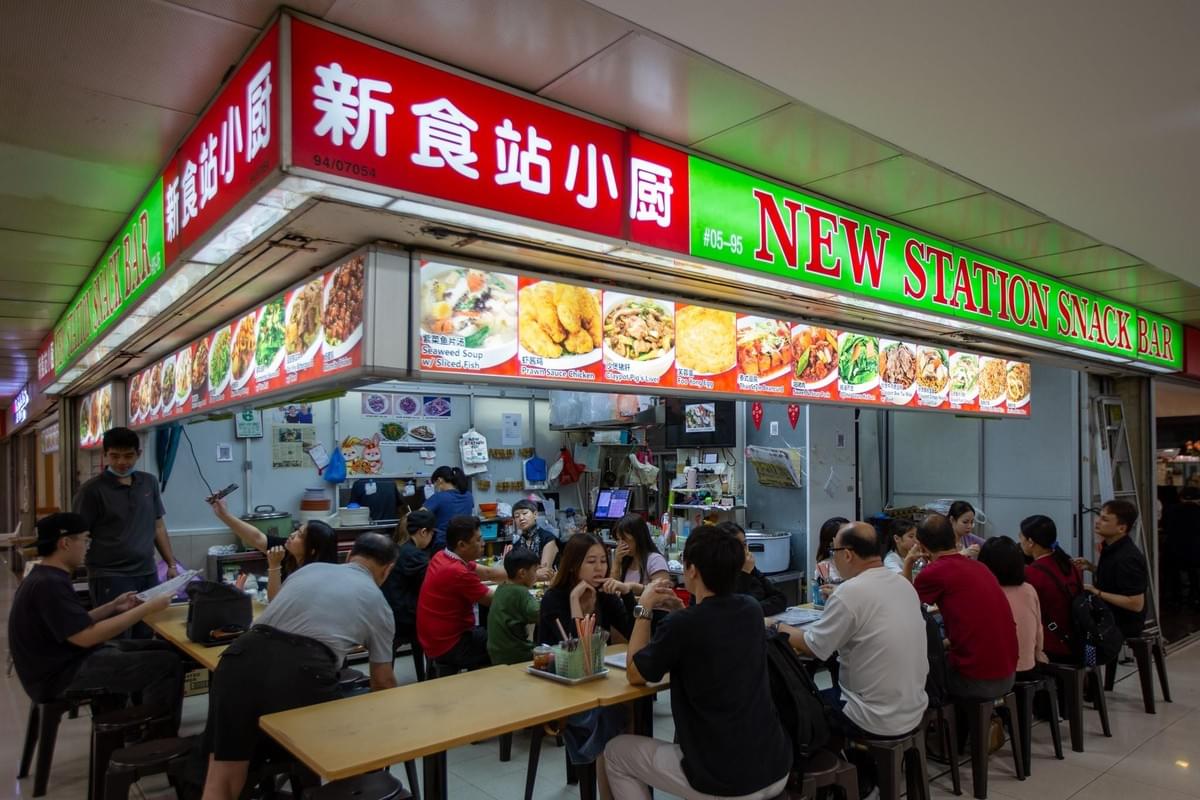 Patrons sit at long communal tables in front of a brightly lit food stall named "New Station Snack Bar." The stall's glowing red and green signage displays a wide variety of illustrated dishes available for order.