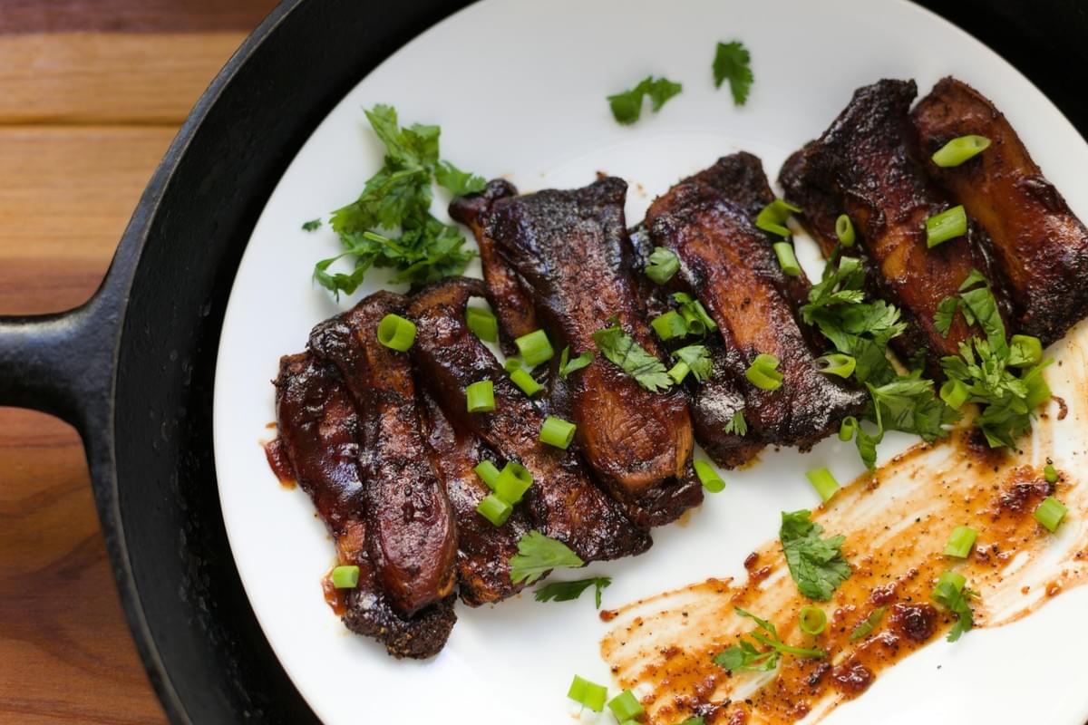 Pan-seared, dark-glazed strips of food, possibly mushrooms or meat, are arranged on a white plate inside a cast-iron skillet. They are garnished with bright green chopped scallions and fresh cilantro, with a decorative smear of savory sauce on the side.