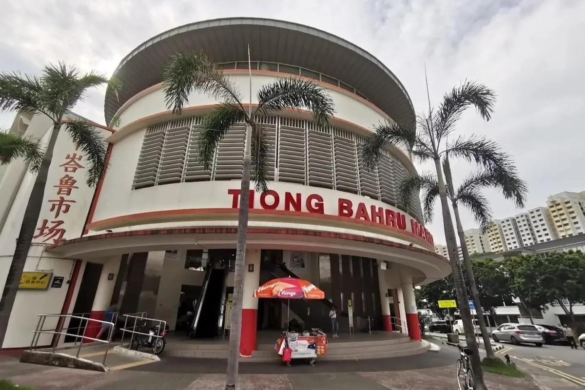 This image shows the circular, multi-story facade of the Tiong Bahru Market in Singapore, featuring its distinctive red lettering and Art Deco-inspired architecture. Palm trees and a small food cart stand in the foreground, while modern residential blocks and parked cars line the street in the background.