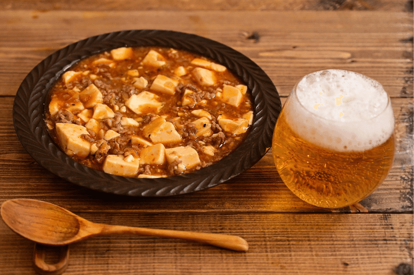 A top-down view of Mapo Tofu served in a dark, textured rimmed plate on a rustic wooden table, accompanied by a wooden spoon and a glass of foamy beer.