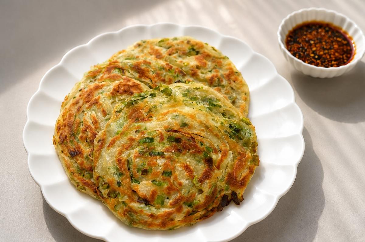 Golden-brown scallion pancakes stacked on a white scalloped plate with a side of dipping sauce.