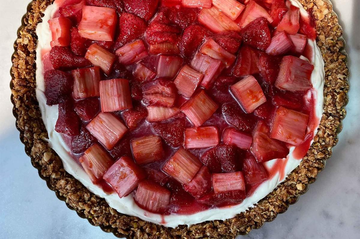 A close-up view of a baked tart or cheesecake topped with a creamy white filling and a thick layer of cooked strawberries and pieces of rhubarb.