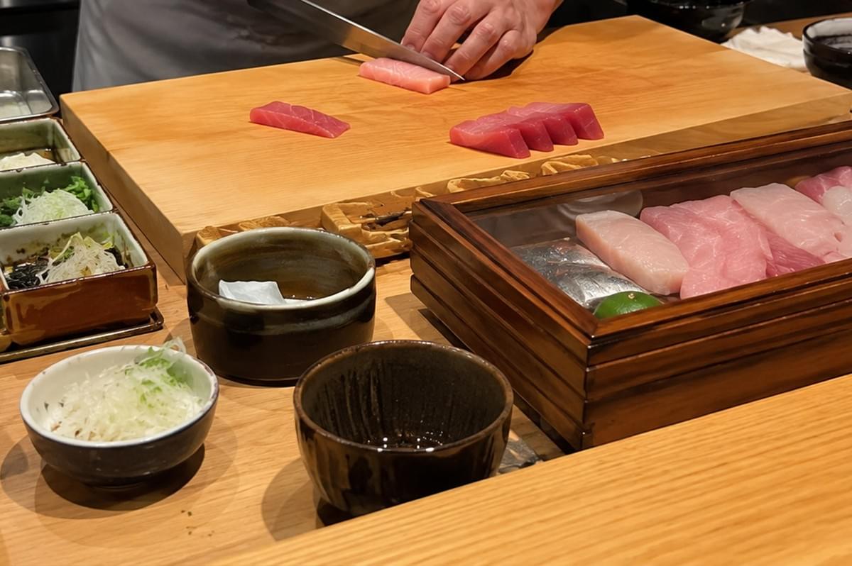 A sushi chef in a white jacket carefully slices pale pink tuna sashimi with a long Yanagiba knife on a clean wooden cutting board, where four fresh red tuna slices already rest. Surrounding the workspace on the light-wood prep counter are various ceramic side dishes containing garnishes like shredded daikon radish and microgreens, a dark ceramic sauce cup, and an open wooden display box filled with an array of neatly arranged raw fish cuts ranging from white fish to marbled pink otoro.