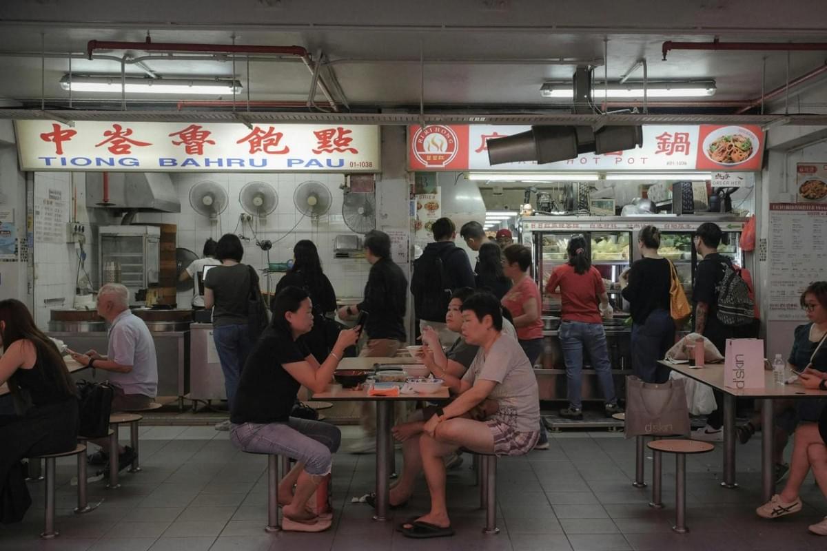 This image captures a bustling scene inside the Tiong Bahru Market hawker center, where patrons are dining at communal tables in front of food stalls like Tiong Bahru Pau. The lively atmosphere is defined by customers queuing for meals and others engaged in conversation over their food under industrial lighting and ceiling fans.