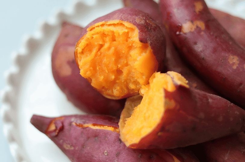 A close-up of a steamed sweet potato broken open on a white plate, revealing a vibrant, fluffy orange interior and steam-softened purple skin.