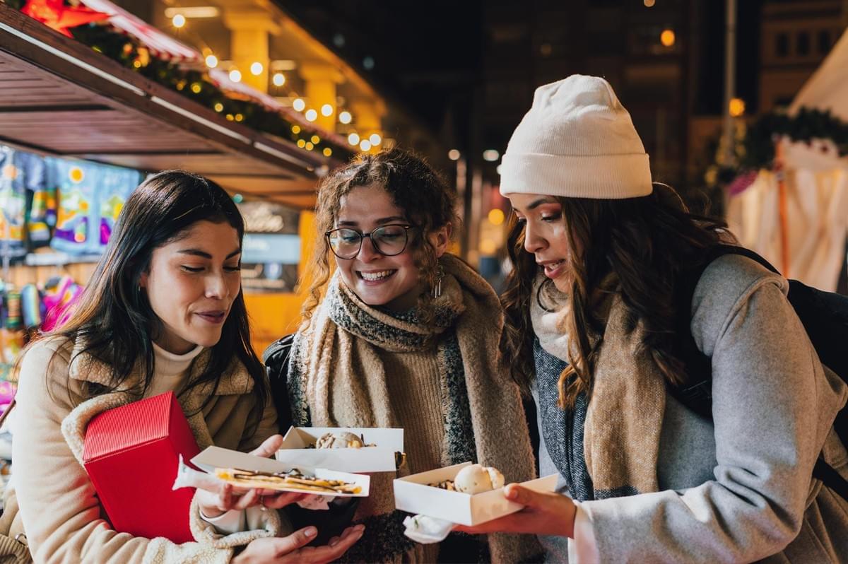 Group of people enjoying street food at an outdoor holiday market, holding festive treats under warm evening lights.
