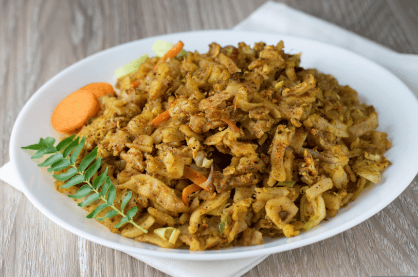 A plate of Kottu Roti featuring visible pieces of egg and spices, garnished with a curry leaf branch and carrot slices on the side.
