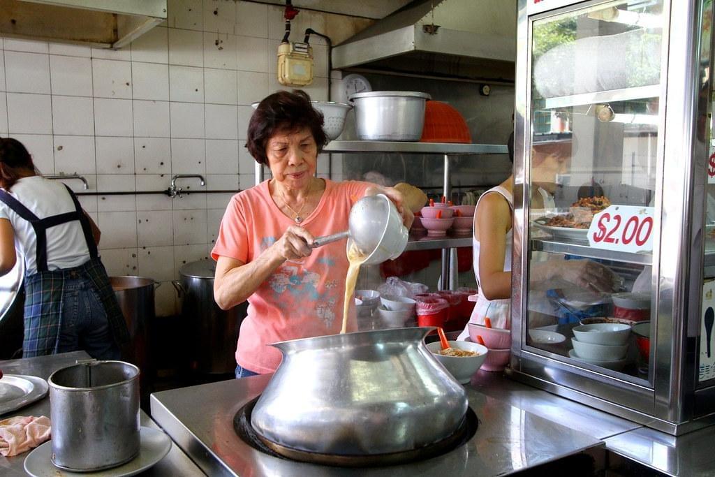 An older woman in a pink shirt pours liquid into a large metal pot in a busy kitchen. She stands between a display case with a price sign and another worker prepping in the background.