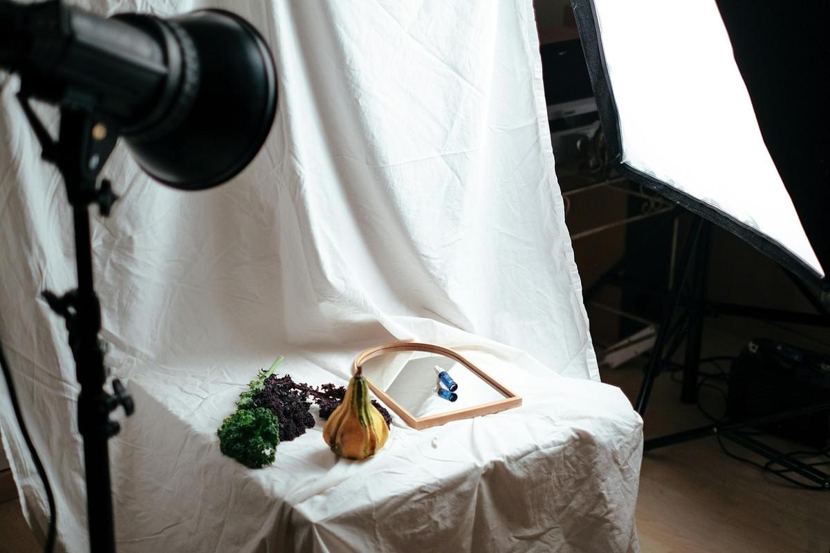 A behind-the-scenes view of a studio photography setup featuring a small product arrangement on a white fabric backdrop. Professional studio lights and a softbox illuminate the setup, which includes a small mirror, a tiny blue dropper bottle, kale leaves, and a decorative gourd.