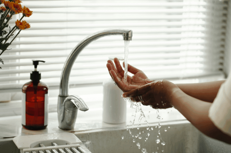 A person thoroughly washing their hands under running water at a kitchen sink with a bottle of soap nearby.