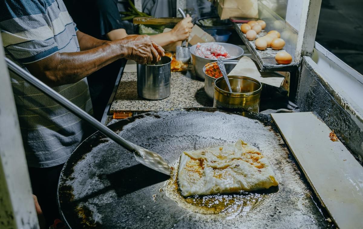 A vendor in a striped shirt uses a metal spatula to fry a square, folded savory pastry on a large, flat, oil-slicked griddle. The workspace behind the cooking surface is lined with fresh brown eggs and metal canisters used for food preparation.