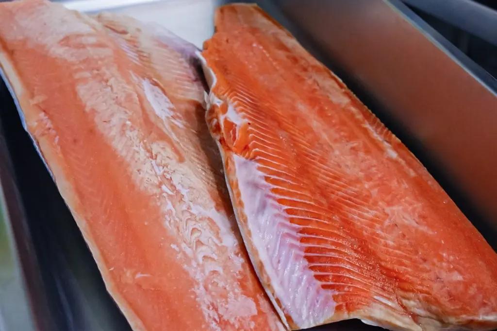 Two large, fresh salmon fillets lie side by side inside a metal tray, showing their vibrant orange flesh and fine white marbling. The clean cuts highlight the fish's smooth texture and rich color, ready for preparation.