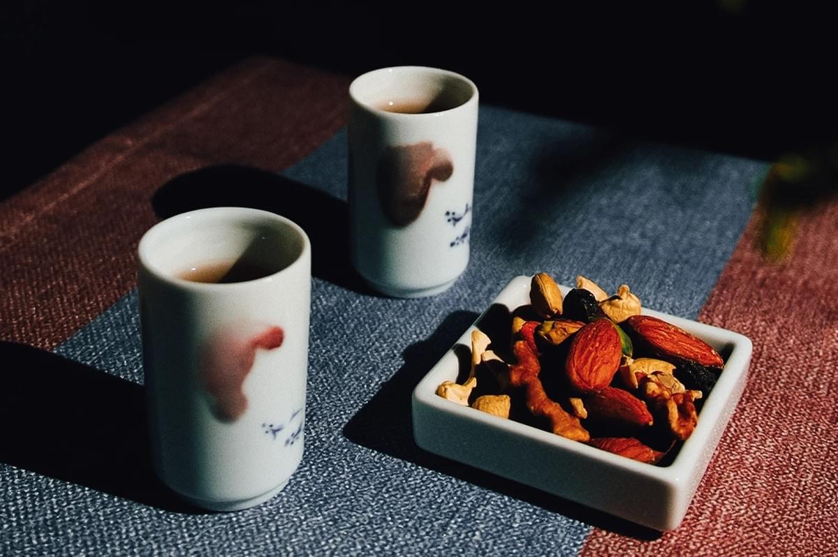 Side‑angle still life shot of two porcelain tea cups beside a small dish of mixed nuts on a textured table, illuminated by dramatic natural light for a calm tea‑pairing scene.
