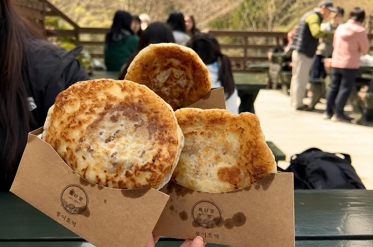 Three hotteok (Korean sweet pancakes) held up in paper sleeves outdoors with people blurred in the background.