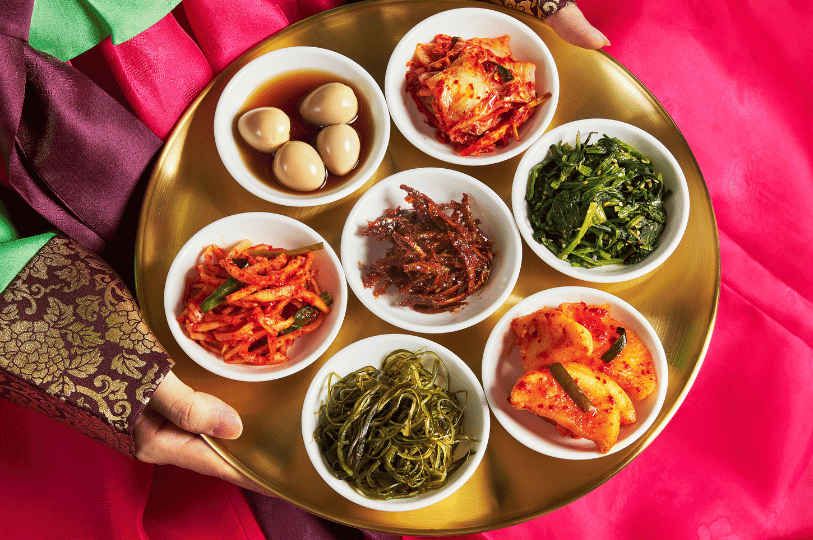 A person wearing a vibrant traditional Korean hanbok holds a round brass tray presenting seven small white bowls of assorted banchan, including soy-marinated quail eggs, classic cabbage kimchi, seasoned spinach, and stir-fried anchovies.