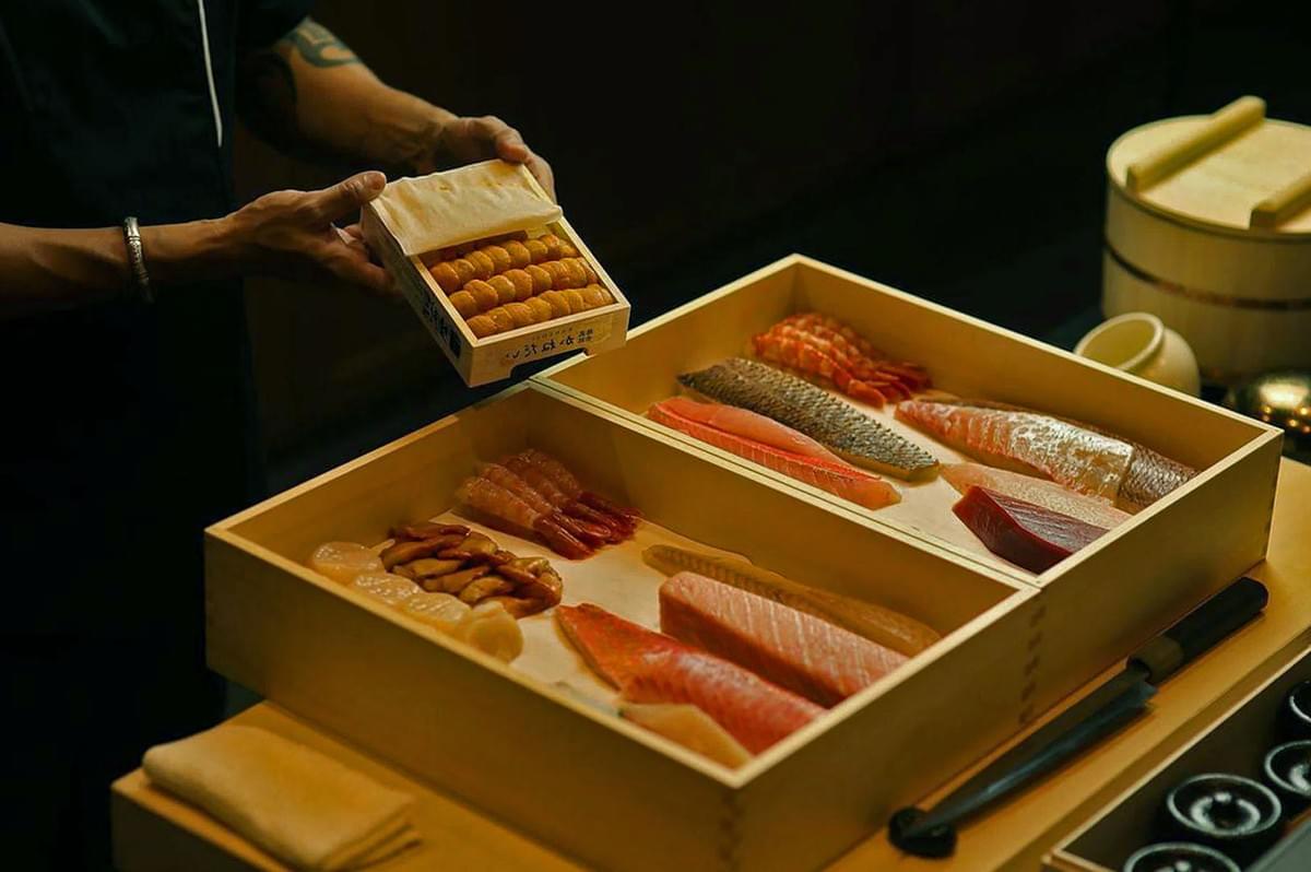A person holding a box of uni over a wooden platter filled with various sliced sushi-grade fish, including salmon and tuna, in a dimly lit setting.