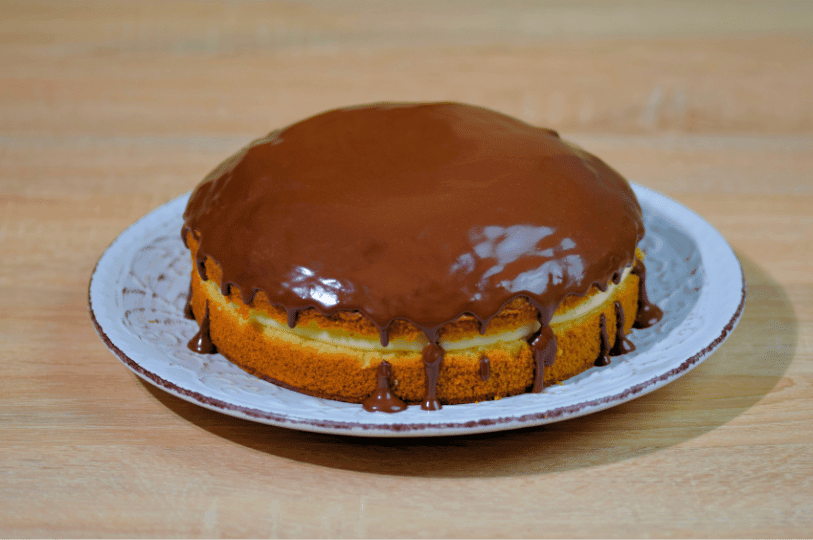 A full view of a homemade Boston Cream Pie on a white ceramic plate, featuring a smooth, dark chocolate ganache glaze with decorative drips over the edges of the cake.