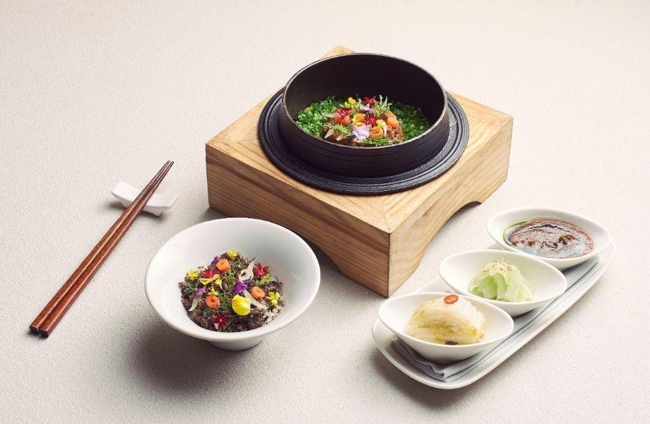 An elegantly plated meal features a cast-iron rice pot set in a wooden base, filled with seasoned rice and colorful edible flower garnishes. It is served alongside a white bowl with a matching dish, three small condiment dishes containing kimchi and sauces, and a pair of wooden chopsticks.