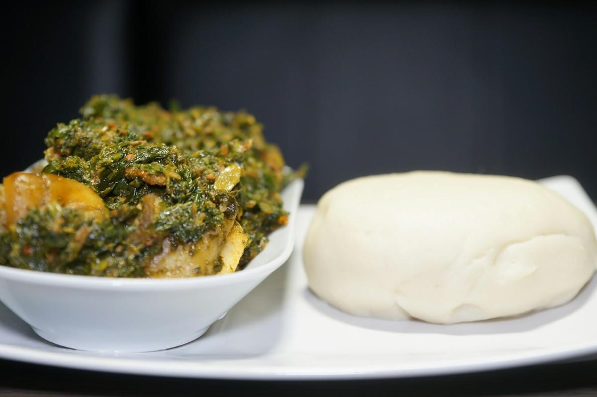 A white bowl of Egusi soup on a plate next to a plate of white bread, representing Nigerian culinary techniques.