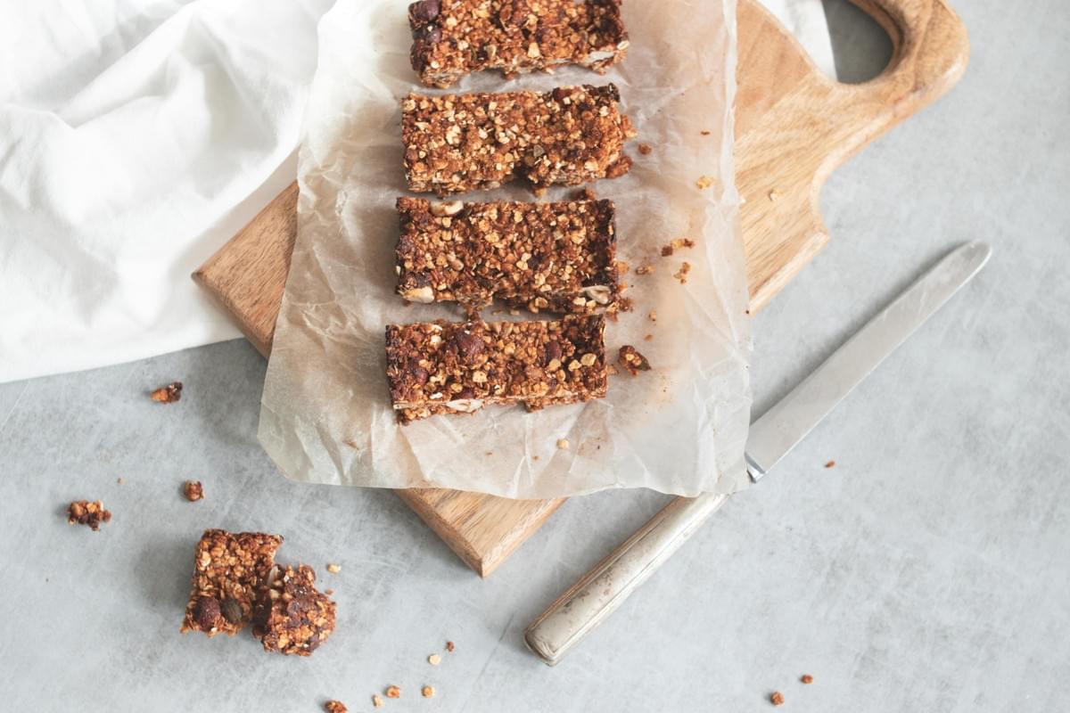 Four dark, textured granola bars are arranged on crinkled parchment paper atop a wooden cutting board. Scattered crumbs and a vintage silver knife rest on the cool grey surface, framing the rustic, homemade snack.