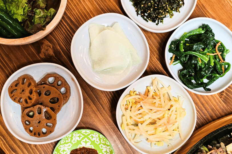 A top-down, close-up view of a wooden table set with several small white bowls of everyday Korean side dishes, highlighting sweet braised lotus root, translucent pickled radish slices, seasoned bean sprouts, and bright green spinach namul.