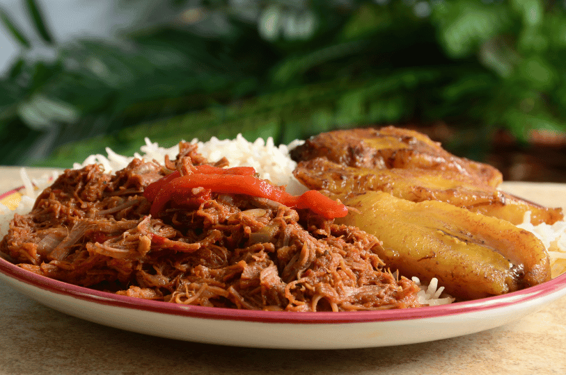A classic Cuban presentation of Ropa Vieja featuring a heap of tender, shredded beef in a savory tomato-based sauce, served alongside white rice and sweet fried plantains (maduros).