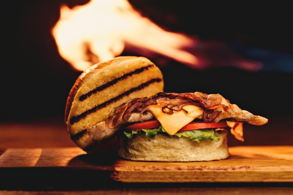 A gourmet chicken sandwich is shown open-faced on a wooden board, featuring a grilled chicken breast, melted cheese, bacon, lettuce, tomato, and a toasted bun. In the background, dramatic flames erupt from a grill, creating a warm and fiery atmosphere.