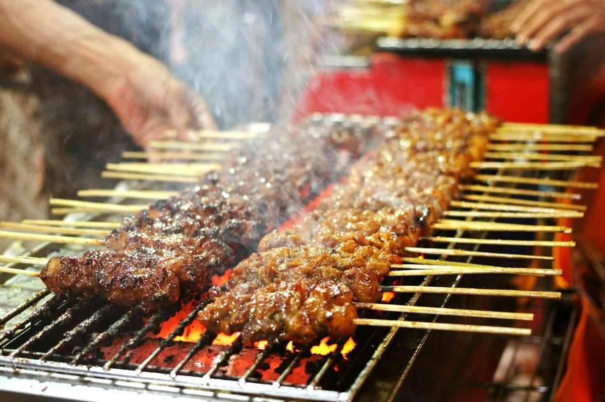 A chef cooking skewers of meat on a grill, preparing traditional Singapore Satay, with smoke rising in the background.