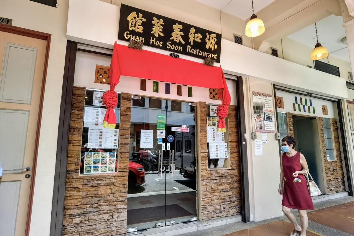 The entrance of Guan Hoe Soon Restaurant is framed by textured stone pillars and a festive red banner draped over the glass doors. A woman wearing a face mask and a red dress walks along the tiled pavement in front of the establishment, which displays several menus and signs in its windows.