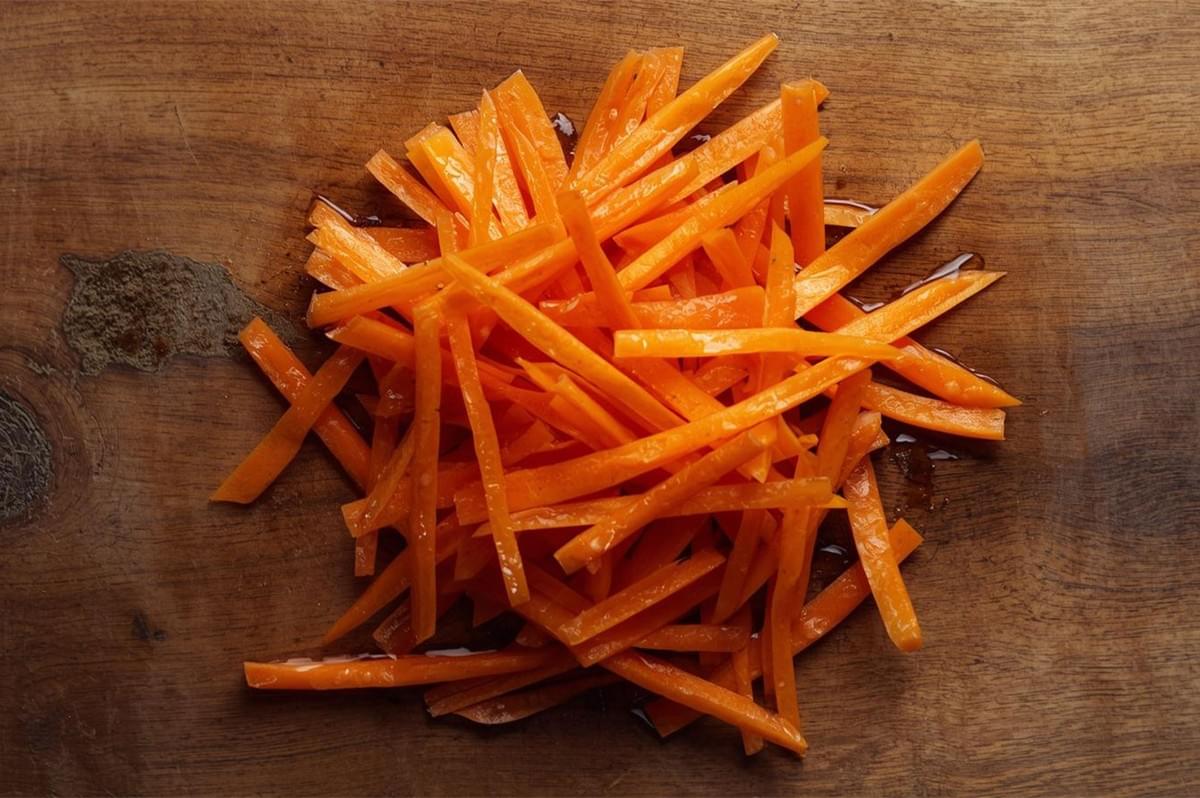 An overhead shot of a scattered pile of bright orange carrots cut into thin, matchstick shapes, demonstrating the julienne cut technique on a dark wooden board.