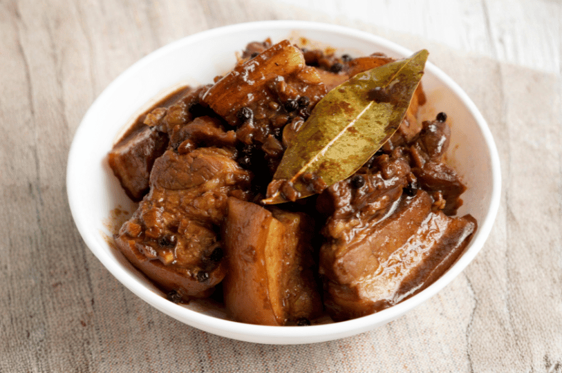 A top-down view of a white bowl containing Filipino Adobo, highlighting the rich, dark brown braising liquid and a prominent dried bay leaf resting on the meat.