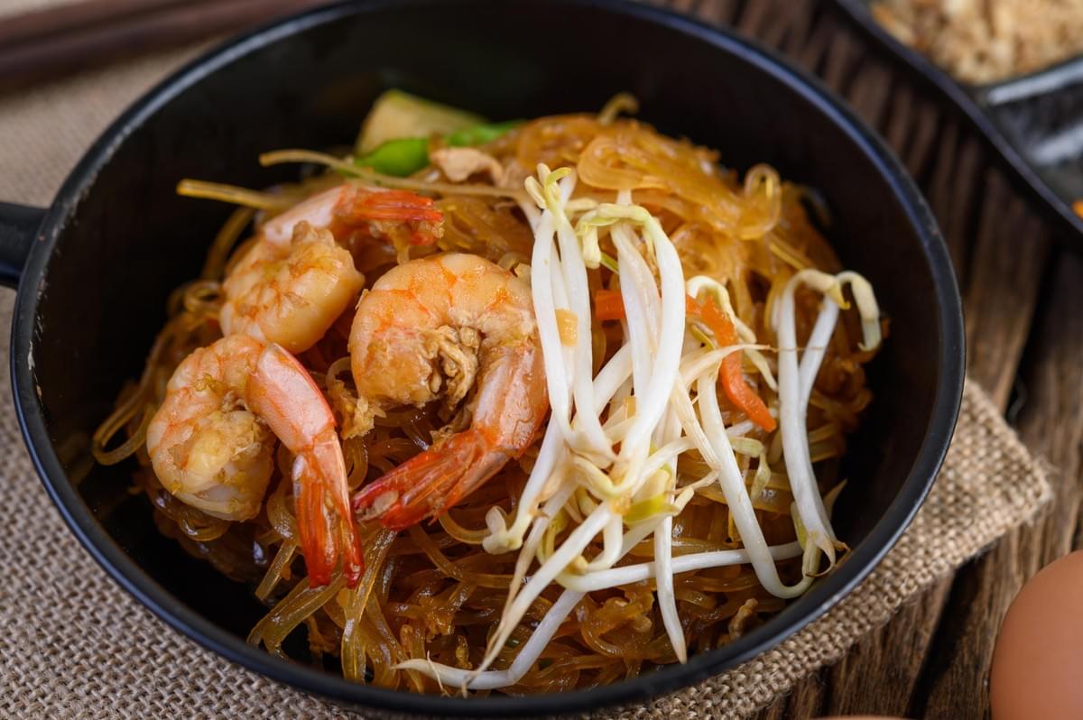 The image shows a close-up view of a serving of Pad Thai with thin, glass-like noodles in a black bowl. The dish is topped with several cooked, orange shrimp and a handful of fresh white bean sprouts.