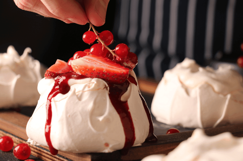 A chef’s hand placing a sprig of red currants onto a mini pavlova drizzled with vibrant red berry syrup.